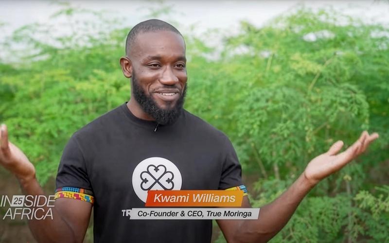 Kwami Williams, Co-Founder and CEO of True Moringa, speaks on CNN’s Inside Africa program. He smiles with arms raised slightly, standing outdoors in front of lush green moringa plants.